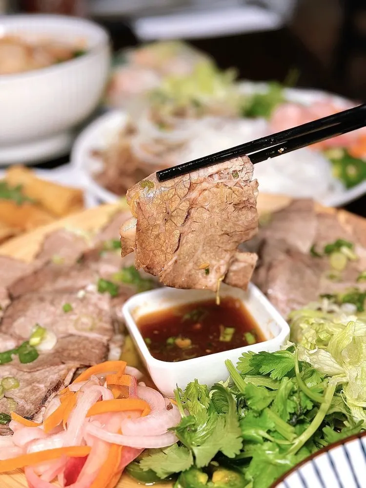 Chopsticks lifting slow-cooked brisket over a dipping sauce, with pickled carrots and herbs