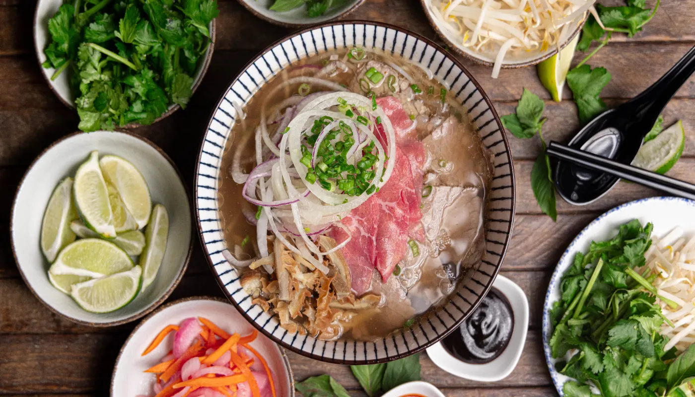 Overhead view of the signature phở in a blue pinstriped bowl with rare steak, brisket, tripe, and sliced onions, surrounded by herbs, limes, bean sprouts, and pickled onions