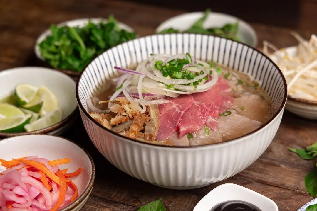 Signature phở in a white ribbed bowl with rare steak, brisket, and tripe, beside limes, herbs, and pickled onions