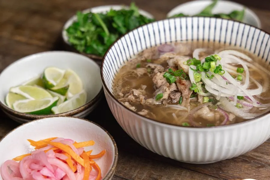 Smoky beef belly phở in a blue pinstriped bowl with onions and scallions, beside limes, herbs, and pickled onions