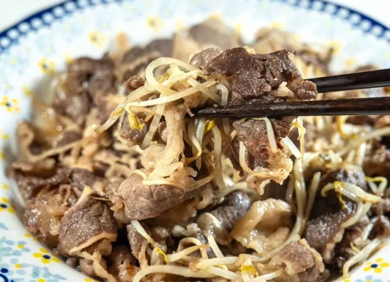 Chopsticks lifting stir-fried beef belly with bean sprouts from a patterned plate