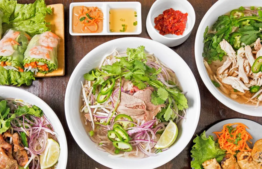 A table of chicken phở, summer rolls, and grilled meats with dipping sauces and herbs